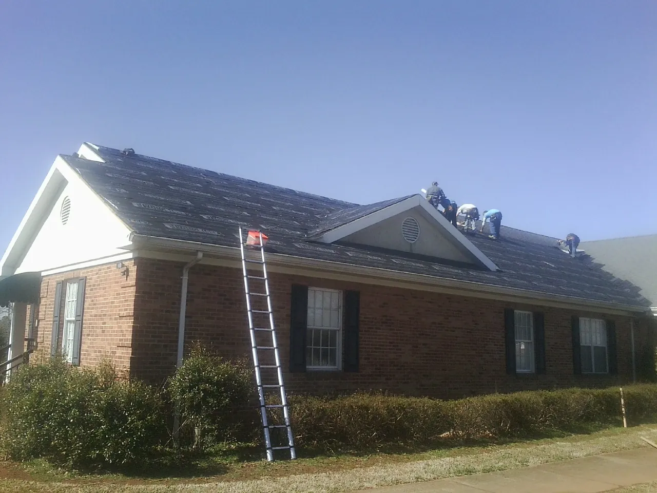 Crew tearing shingles off a brick ranch roof with a ladder set — Metal Roof Repair work by our crew in Nesmith